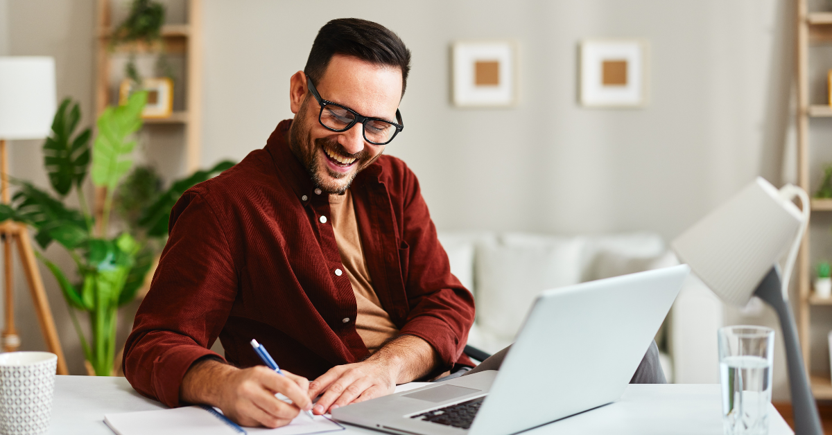 Positieve man met bril en rood blousje zit achter laptop en schrijft iets in schriftje